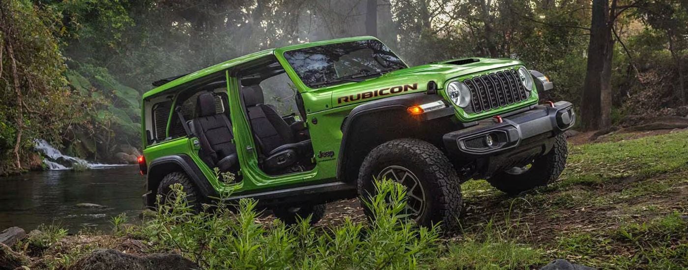 Green 2026 Jeep Wrangler driving near a river in the forest.
